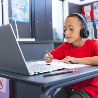 A young student sitting at a desk working in a notebook listening to headphones while looking at a laptop computer.