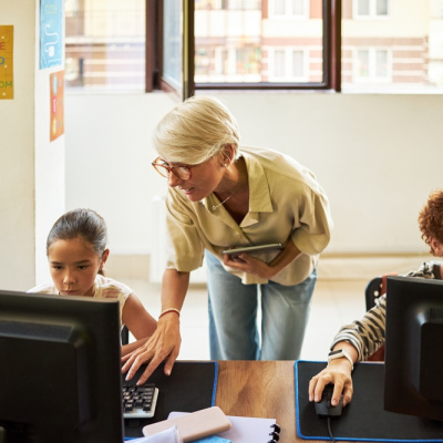 Middle aged educator assisting two young students, working on desktop computers in a classroom.