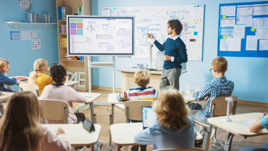 Elementary school science teacher uses interactive digital whiteboard to show classroom full of children how software programming works for robotics.