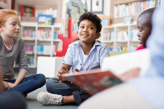 A group of children sit on a floor in circle around a teacher in a school library and listen to a story.