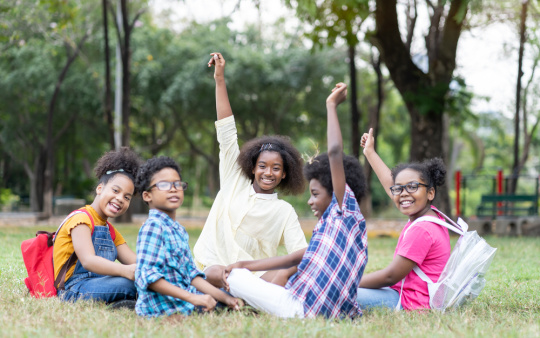 A cheerful group of students sit outside the classroom on the grass, discussing a school project.