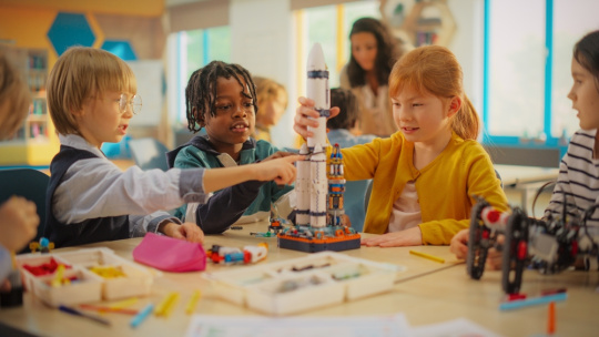 Elementary school students sitting behind a table in a group, building a space rocket model for a science and technology fair.