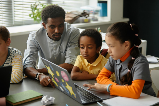 African American male teacher helping younger multiethnic students with using laptop explaining homework while sitting with kids at desk in modern classroom