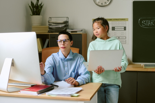 Middle aged educator sitting at desk using desktop computer while a student stands beside her holding laptop, both focusing on computer screen in a classroom.