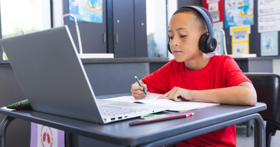 A young student sitting at a desk working in a notebook listening to headphones while looking at a laptop computer.
