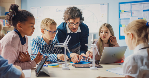 Enthusiastic teacher holding digital tablet explains to young children how wind turbines work.