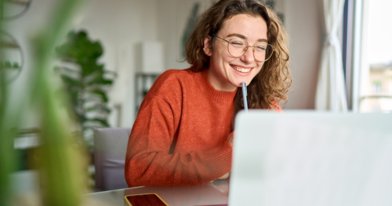 A happy young student sits at her desk writing notes while participating in an online webinar.
