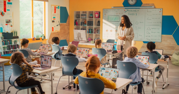 Elementary school children learning basic computer skills with young female teacher at front of class.