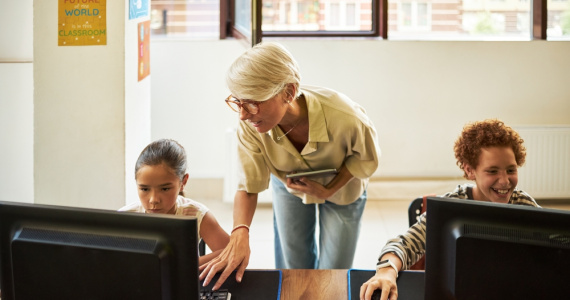 Middle aged educator assisting two young students, working on desktop computers in a classroom.