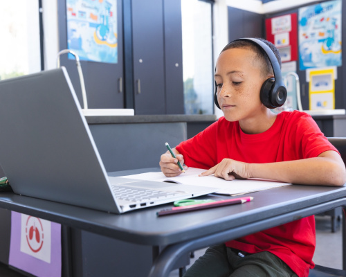 A young student sitting at a desk working in a notebook listening to headphones while looking at a laptop computer.