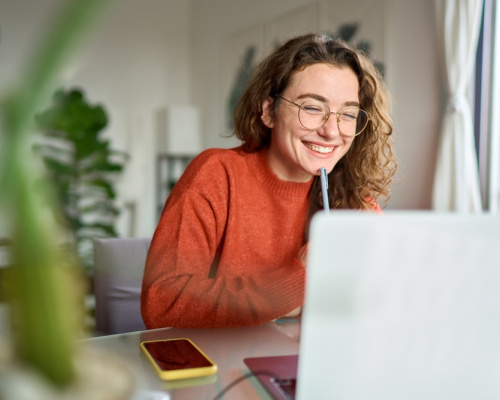 A happy young student sits at her desk writing notes while participating in an online webinar.
