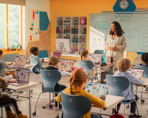 Elementary school children learning basic computer skills with young female teacher at front of class.