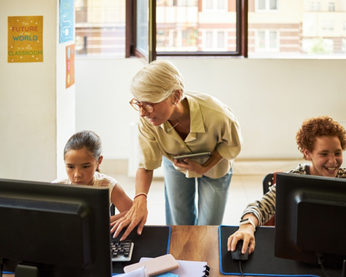 Middle aged educator assisting two young students, working on desktop computers in a classroom.