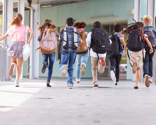 A diverse group of younger students wearing backpacks running to recess.